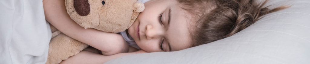 Criança dormindo tranquilamente na cama abraçada a um urso de pelúcia, representando o sono infantil saudável.