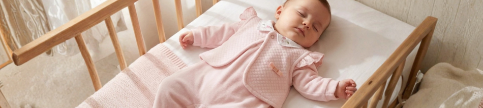 Bebê dormindo tranquilamente no berço de madeira, vestindo um macacão rosa e um colete acolchoado em camadas, ideal para o inverno.