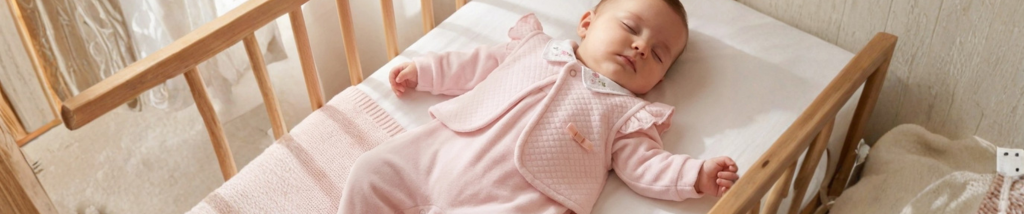 Bebê dormindo tranquilamente no berço de madeira, vestindo um macacão rosa e um colete acolchoado em camadas, ideal para o inverno.